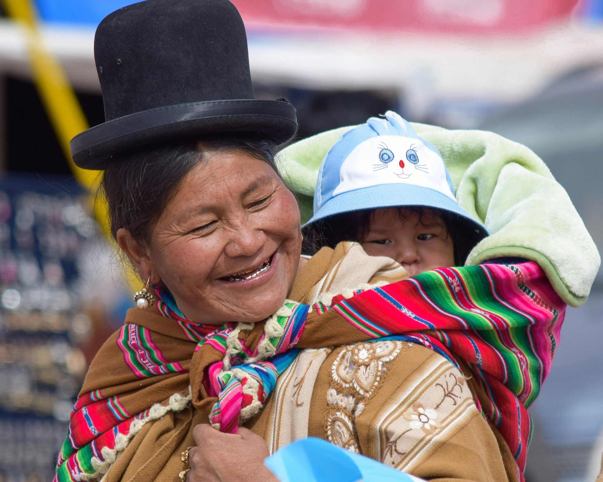A joyful Bolivian woman in traditional clothing carrying a child in a vibrant shawl, outdoors.