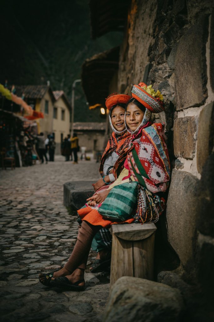 Two women in traditional attire seated on cobblestones in Ollantaytambo, Peru.