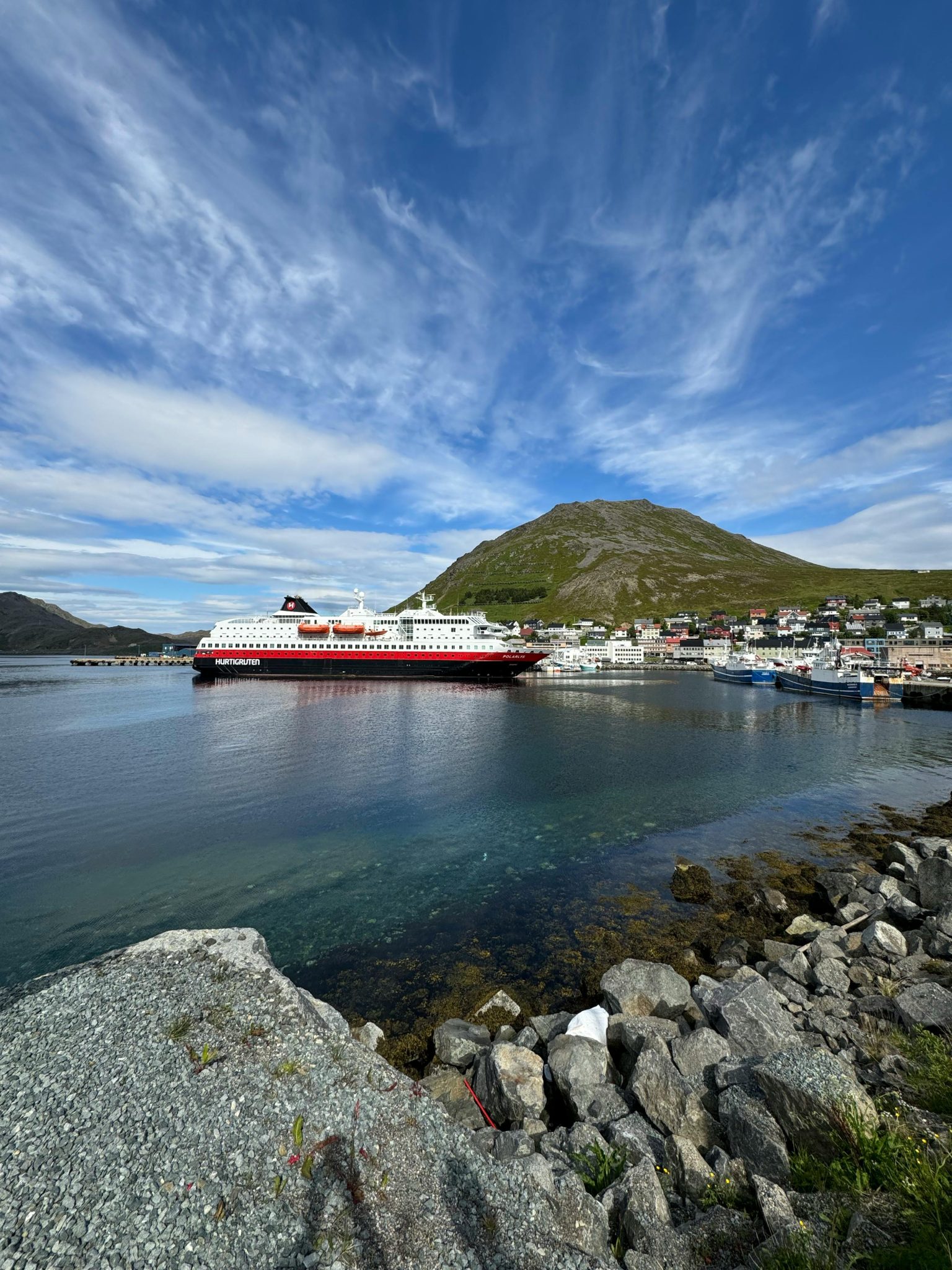 A scenic view of a cruise ship docked in a fjord-side town with a mountain under a bright blue sky.