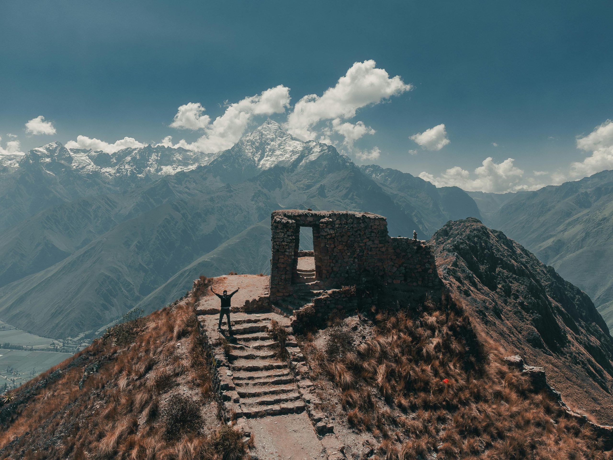 Man raising arms atop Intipunku with snow-capped Andes in Ollantaytambo, Peru.
